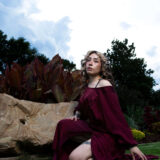 Woman wearing a flowing burgundy dress posing beside decorative stonework and garden foliage.