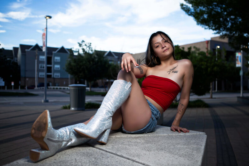 Fashion portrait of a model seated on a concrete bench in downtown Oklahoma City during blue hour, wearing sparkling silver boots and a red top.