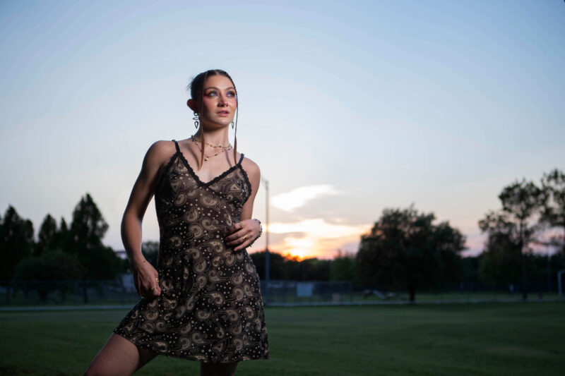 Portrait of a woman wearing a patterned black dress standing in the grass at Ray Trent Park in Del City during sunset, illuminated with off-camera lighting against a colorful evening sky.