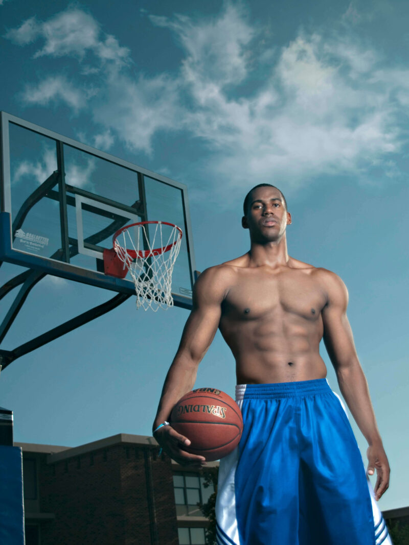 Athletic portrait of a basketball player holding a basketball beneath an outdoor hoop against a dramatic blue sky.