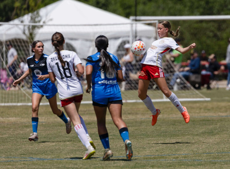 U16 girls soccer player from Oklahoma Energy in white jumps to control the ball while Amarillo Rush defenders in blue close in during the 2026 NOKC Spring Finale