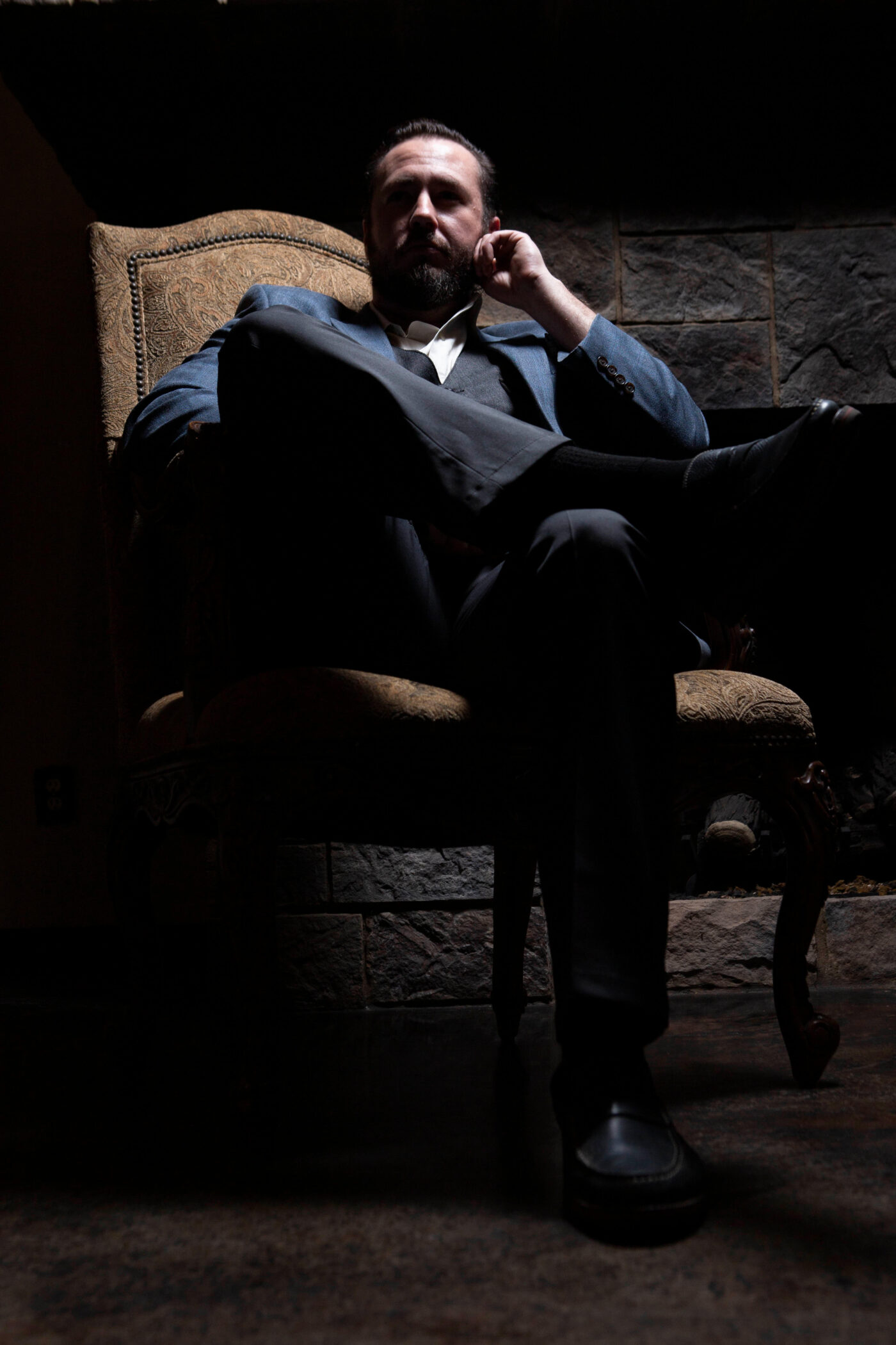 Low-key cinematic portrait of a man in a blue suit seated in an ornate chair beside a stone fireplace during a 1940s-themed photomeet at Castle Falls.