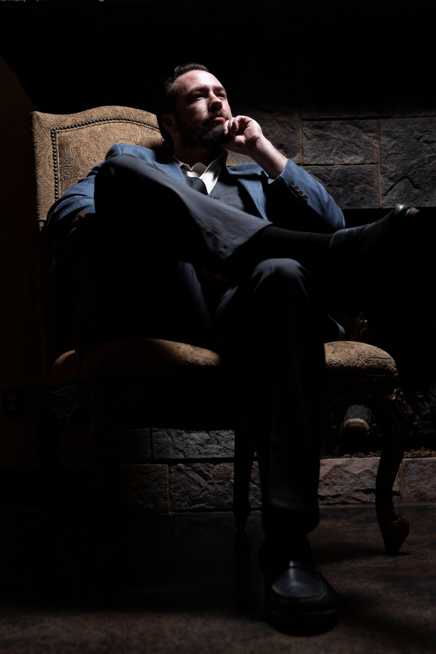 Portrait of a man in a blue suit seated thoughtfully in a vintage chair with dramatic lighting during a 1940s-inspired photography event.