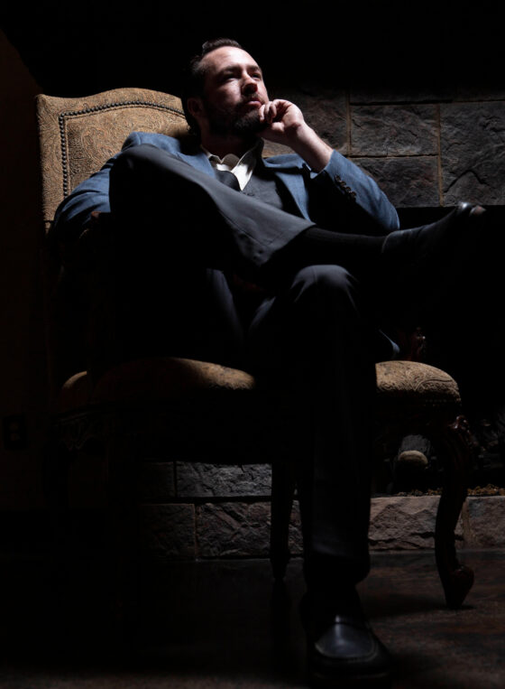 Portrait of a man in a blue suit seated thoughtfully in a vintage chair with dramatic lighting during a 1940s-inspired photography event.