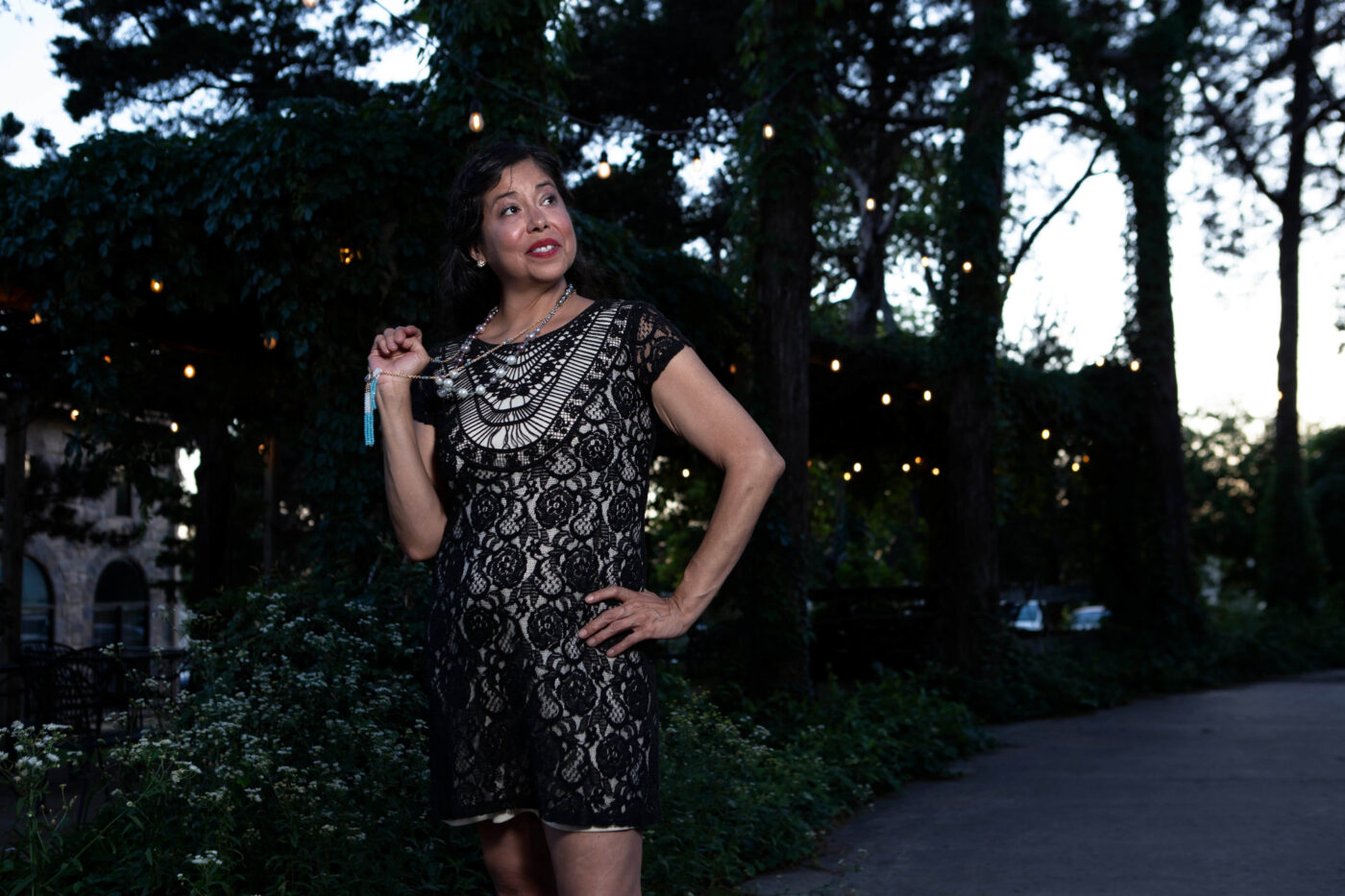 Portrait of a woman in a black lace dress holding a necklace while posing beneath string lights at Castle Falls during a vintage-themed photography event.