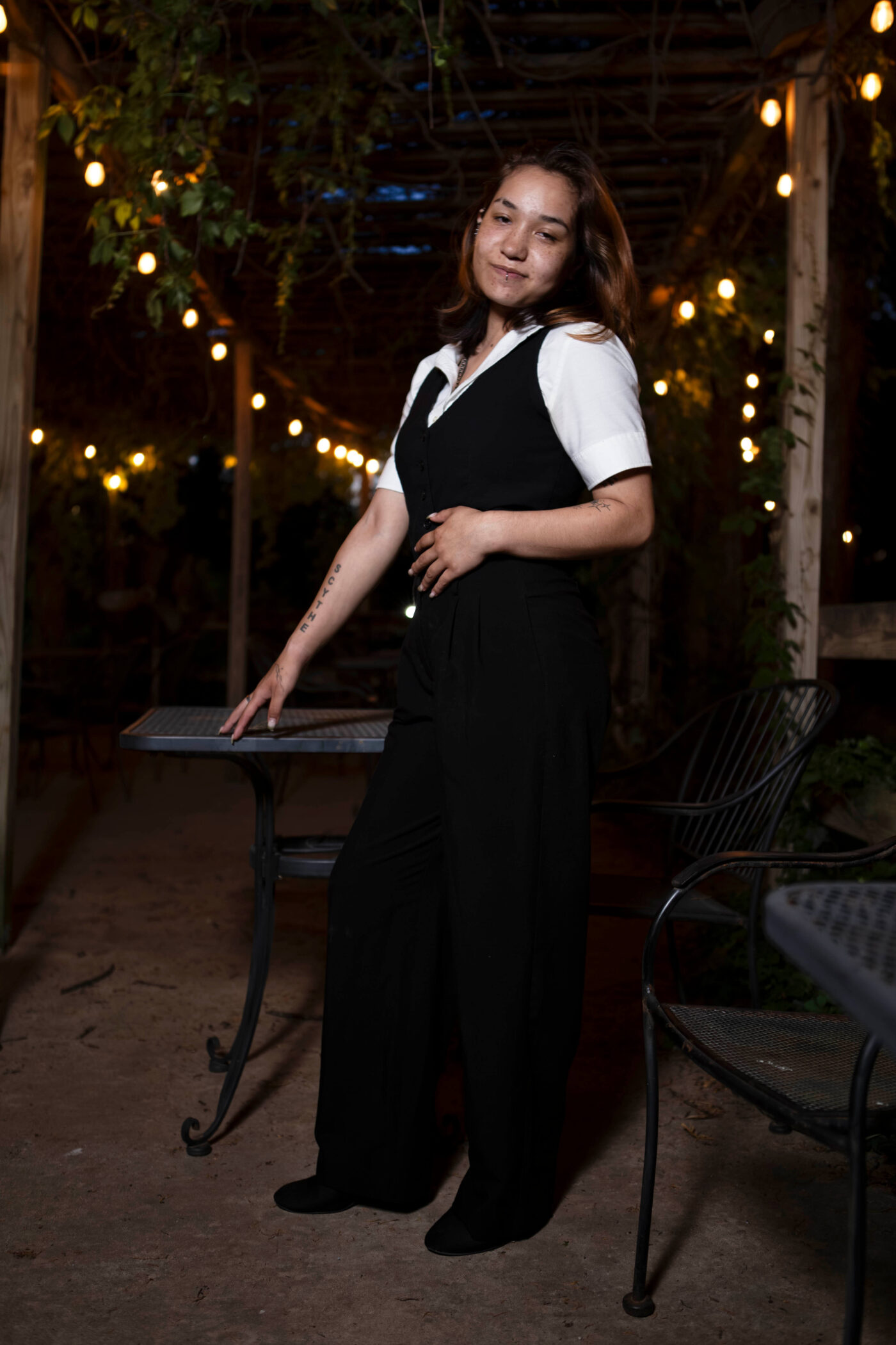 Portrait of model toxicwoman55 standing beneath warm string lights at Castle Falls during a 1940s-themed photomeet, wearing a black vintage-inspired outfit with a white collared shirt.