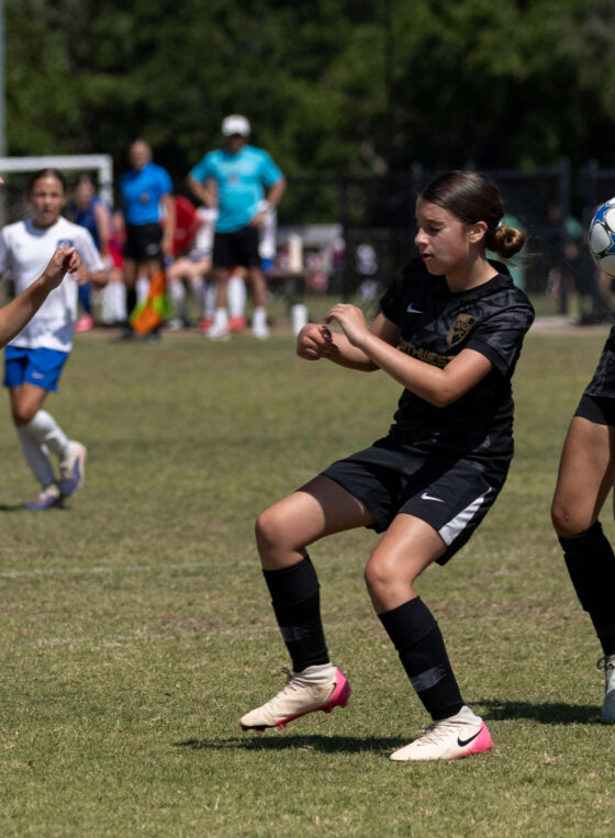 Claremore SC Select player in white closes in defensively as two Northwest Optimist SC players in black attempt to control a bouncing soccer ball during a U13 Girls tournament match in Norman, Oklahoma.