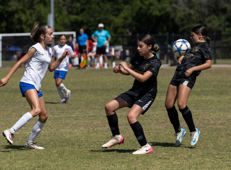 Claremore SC Select player in white closes in defensively as two Northwest Optimist SC players in black attempt to control a bouncing soccer ball during a U13 Girls tournament match in Norman, Oklahoma.