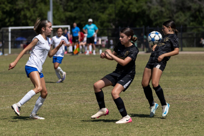 Claremore SC Select player in white closes in defensively as two Northwest Optimist SC players in black attempt to control a bouncing soccer ball during a U13 Girls tournament match in Norman, Oklahoma.