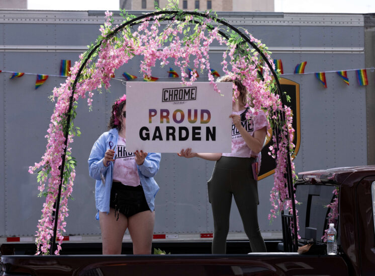 Parade participants stand beneath a flower-covered arch holding a Proud Garden sign.