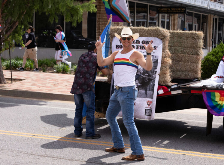 Parade participant wearing a rainbow tank top poses in front of a rodeo-themed float.