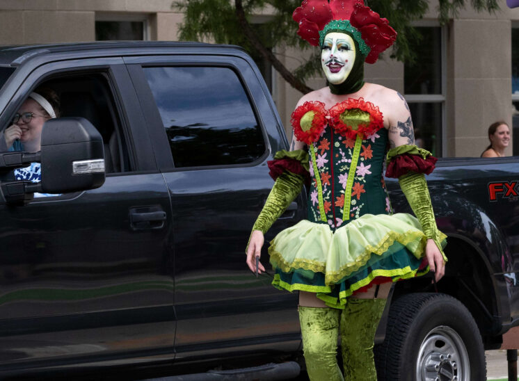 Performer wearing an elaborate green floral costume walks beside a parade vehicle.