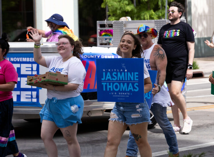 Pride parade participants walk alongside a campaign float carrying signs and waving to spectators.