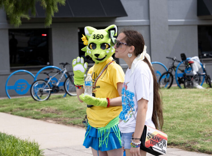 A costumed attendee wearing a bright green mascot-style head poses beside another participant at the Pride festival.