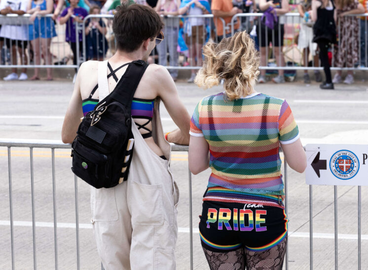 Two spectators wearing rainbow-themed clothing stand behind a barricade watching the Pride parade.