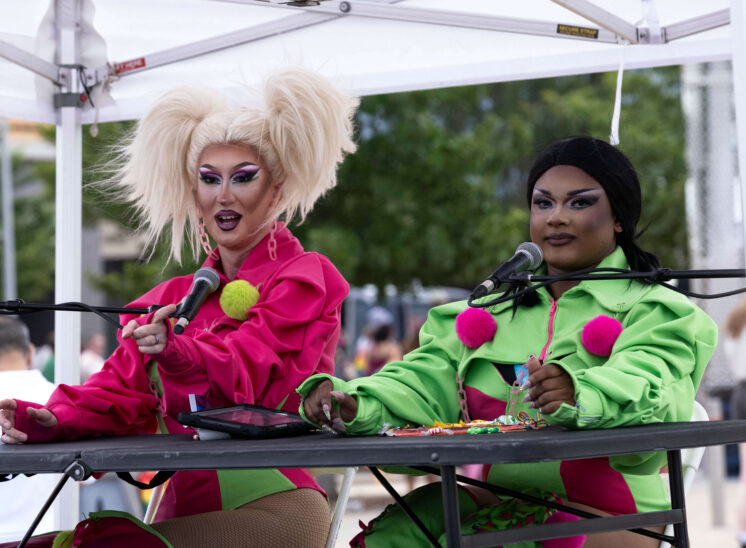 Two brightly dressed presenters sit behind microphones during a Pride festival stage event.