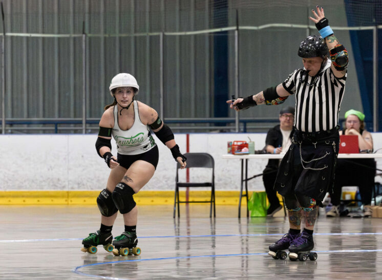 Capital City Crushers jammer skates forward while a referee signals during a roller derby match at Arctic Edge Ice Arena.