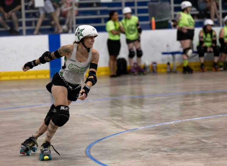 Capital City Crushers skater rounds the track during a roller derby bout at Arctic Edge Ice Arena while Outlaws skaters watch from the bench.