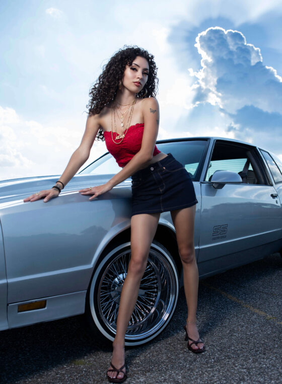 Jas poses beside a silver Chevrolet Monte Carlo SS on a rural roadway, wearing a red lace top and black skirt while dramatic storm clouds and sun rays illuminate the sky behind her.
