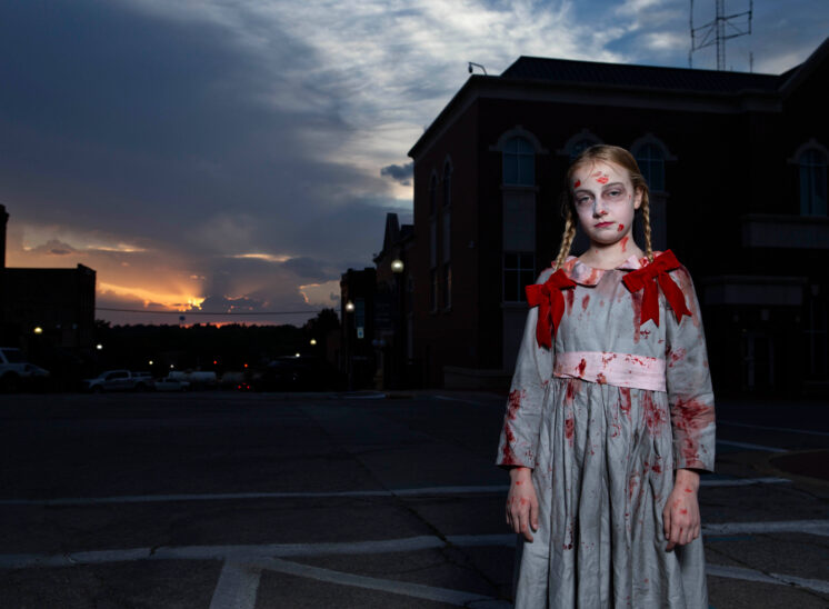 Young Halloween participant dressed as a ghostly Victorian-style doll, wearing a pale blue bloodstained dress with red shoulder bows and braided hair, standing in downtown Guthrie at sunset.