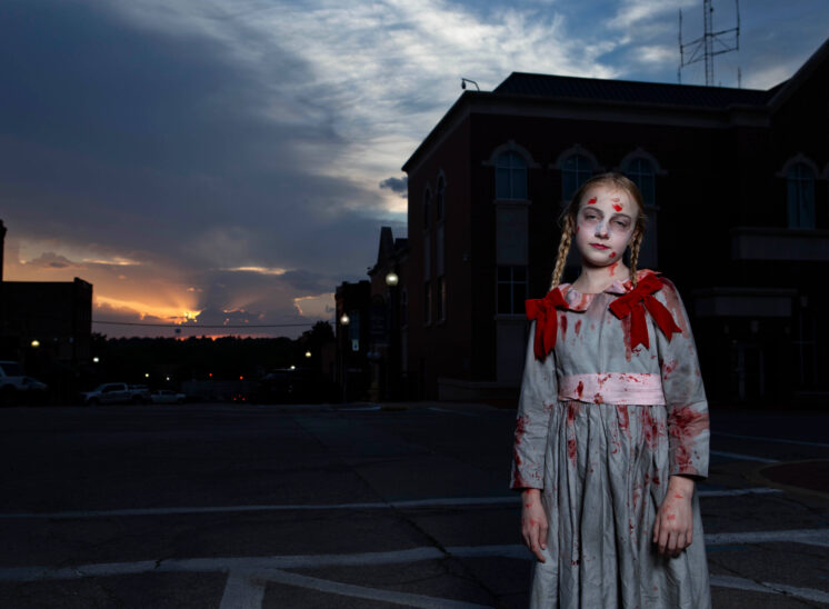 Young girl in a bloodstained vintage-style Halloween dress with red bows and braided hair, tilting her head slightly while standing beneath a dramatic evening sky in Guthrie, Oklahoma.