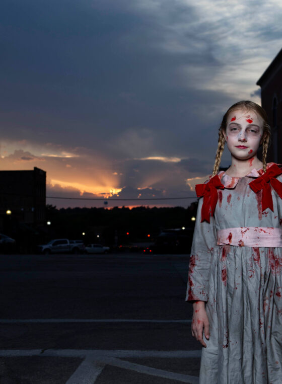 Young Halloween participant with braided hair, pale makeup, and a bloodstained gray dress decorated with red bows, standing in downtown Guthrie against a sunset and historic buildings.