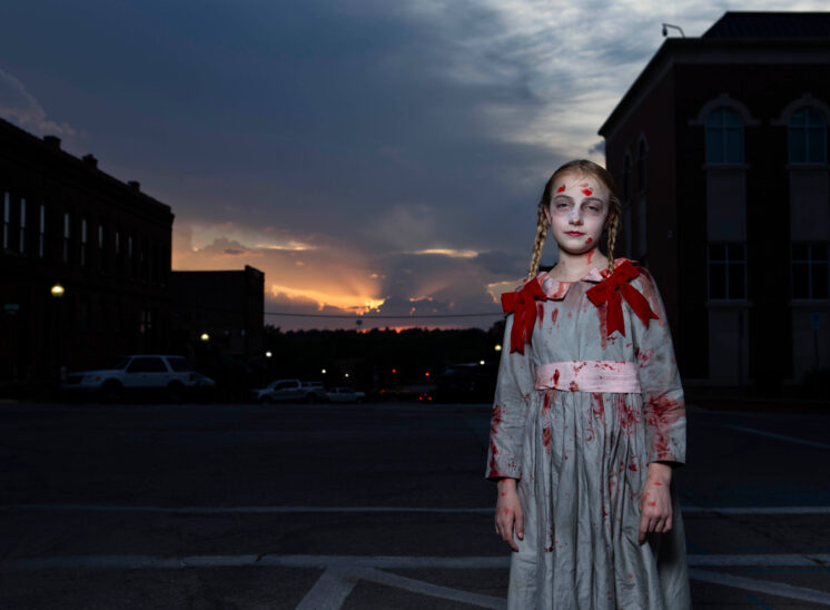 Young Halloween participant with braided hair, pale makeup, and a bloodstained gray dress decorated with red bows, standing in downtown Guthrie against a sunset and historic buildings.