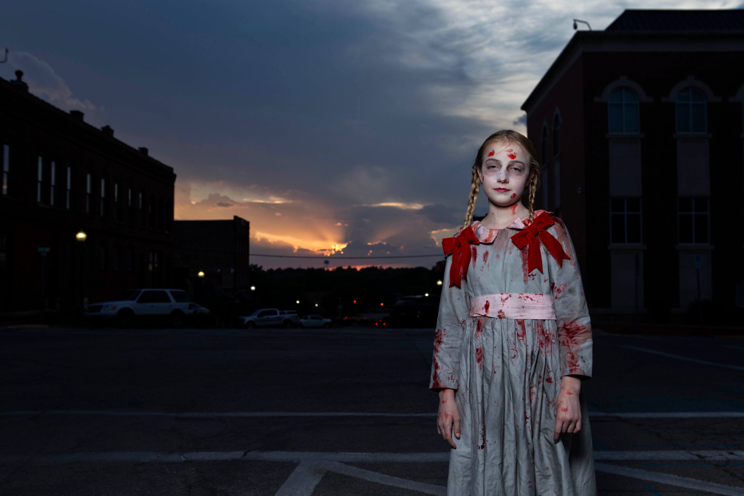 Young Halloween participant with braided hair, pale makeup, and a bloodstained gray dress decorated with red bows, standing in downtown Guthrie against a sunset and historic buildings.