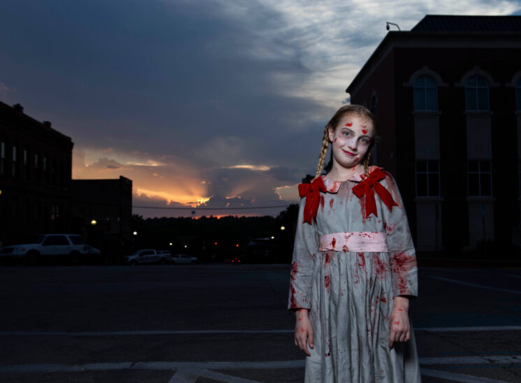 Young girl dressed as a ghostly Halloween doll, wearing a bloodstained pale dress with red shoulder bows and braided hair, smiling slightly while posing in downtown Guthrie at sunset.