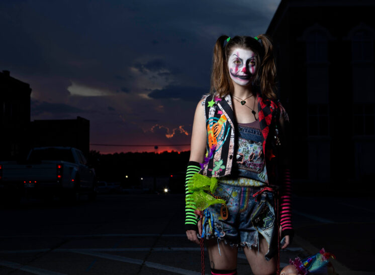 Full-length portrait of a Halloween participant dressed as a sinister clown with white and pink face paint, colorful distressed clothing, striped arm warmers, and pigtails, standing beneath a dark evening sky in Guthrie.