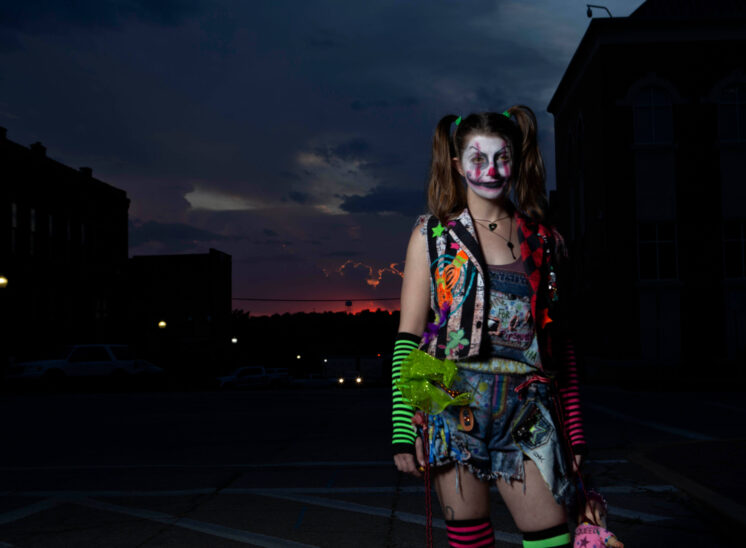 Halloween participant with sinister white clown makeup, brown pigtails, a colorful patchwork vest, striped arm warmers, and distressed denim shorts, posing against a dark Oklahoma sunset.