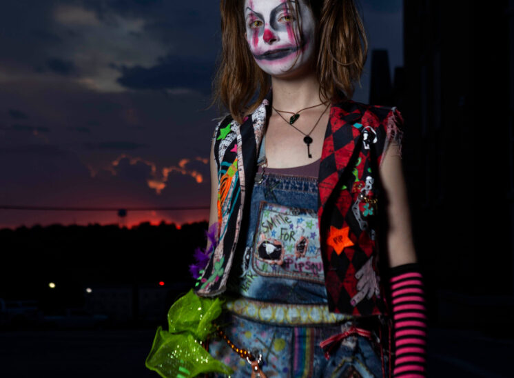Vertical Halloween portrait of a participant wearing sinister clown makeup, brown pigtails, a colorful patchwork outfit, and pink striped arm warmers while holding a colorful doll against a dark twilight sky.