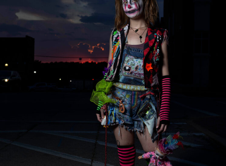 Full-length vertical portrait of a Halloween participant with sinister clown makeup, colorful distressed clothing, striped stockings, black boots, and pigtails, holding a decorated doll in downtown Guthrie at dusk.