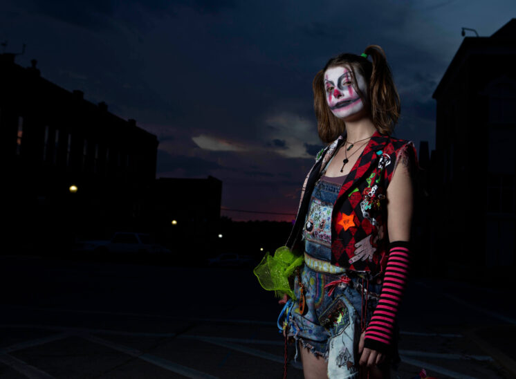 Halloween participant wearing sinister clown makeup, colorful patchwork clothing, and pink striped arm warmers, photographed against the darkening evening sky in downtown Guthrie, Oklahoma.