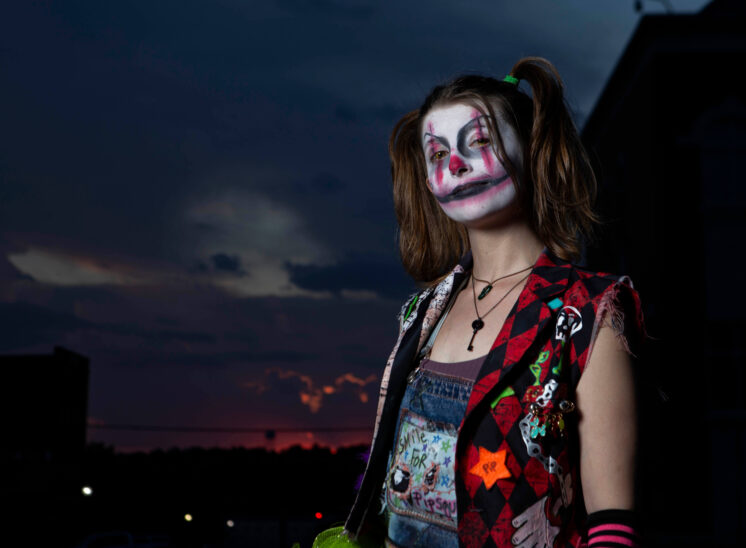 Close-up portrait of a Halloween participant with white clown makeup, exaggerated black and pink smile, brown pigtails, and a colorful patchwork vest against a dramatic twilight sky.