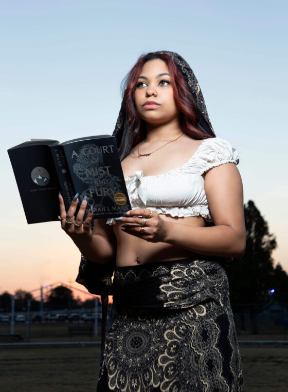 Scarlet Ray poses outdoors at sunset, holding an open copy of A Court of Mist and Fury by Sarah J. Maas. She wears a white cropped blouse, a black patterned headscarf, and a flowing black-and-cream patterned skirt against a pastel evening sky.