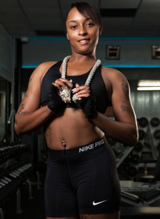 Smiling female fitness model wearing black athletic clothing and holding a thick exercise rope during a professional gym portrait session.