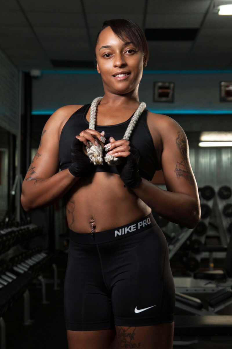 Smiling female fitness model wearing black athletic clothing and holding a thick exercise rope during a professional gym portrait session.