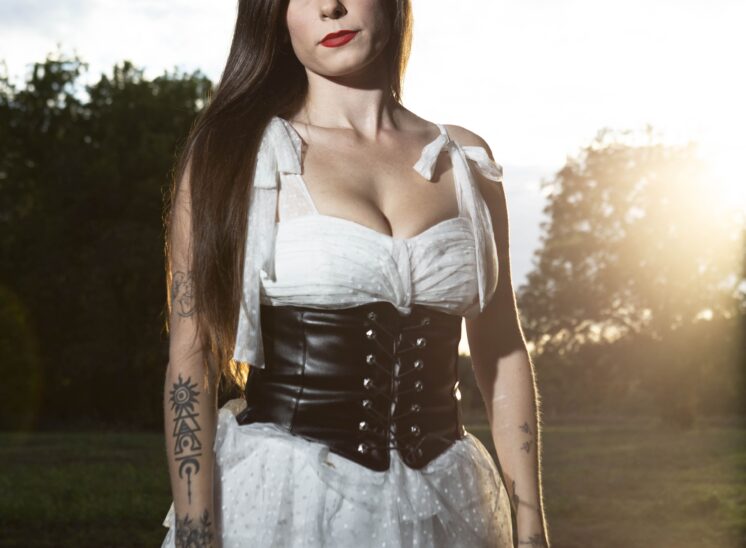 Model @somethingtolivefor222 wearing a white dress and black lace-up corset, posing beside reflective water during sunset at Ray Trent Park.