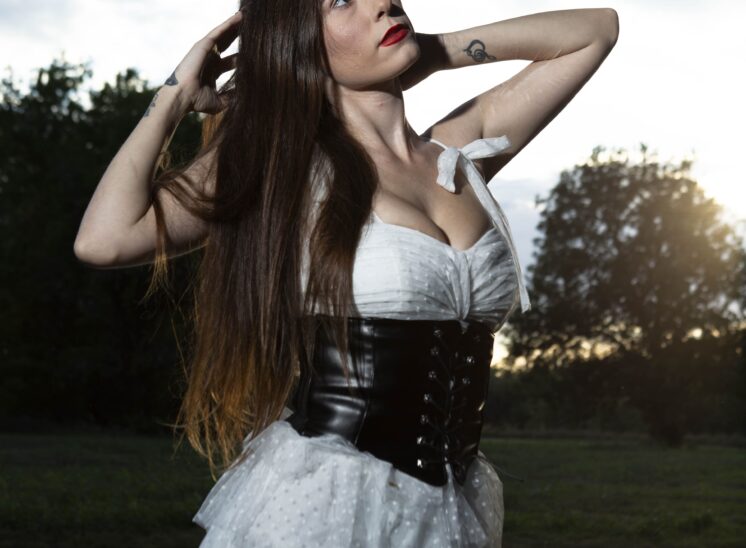 @somethingtolivefor222 wearing a white dress and black corset, raising her arms into her long dark hair against a sunset sky at Ray Trent Park.