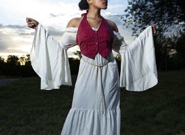 @nq.liyahh wearing a long white dress with flowing sleeves and a burgundy striped vest extends both arms against a blue and gold evening sky.