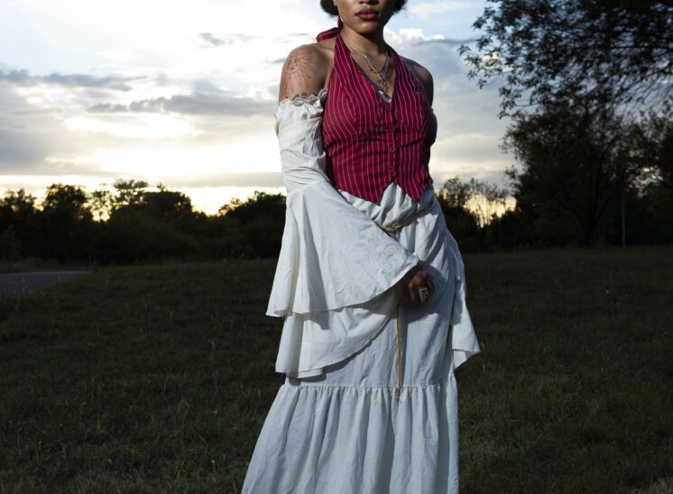 @nq.liyahh wearing a burgundy striped vest over a flowing white dress stands in a grassy field at sunset, looking directly at the camera.