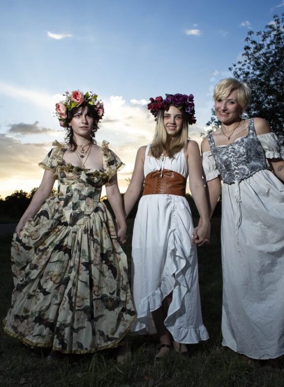 Three models pose together at sunset in Ray Trent Park. @maddenchaotic_cos wears a floral gown and pink flower crown, an unidentified model wears a white dress with a brown corset and purple flower crown, and @eternalhallowcos wears a flowing white and gray dress.