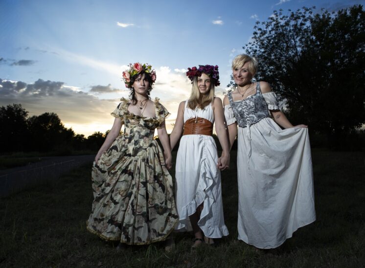 Three models pose together at sunset in Ray Trent Park. @maddenchaotic_cos wears a floral gown and pink flower crown, an unidentified model wears a white dress with a brown corset and purple flower crown, and @eternalhallowcos wears a flowing white and gray dress.