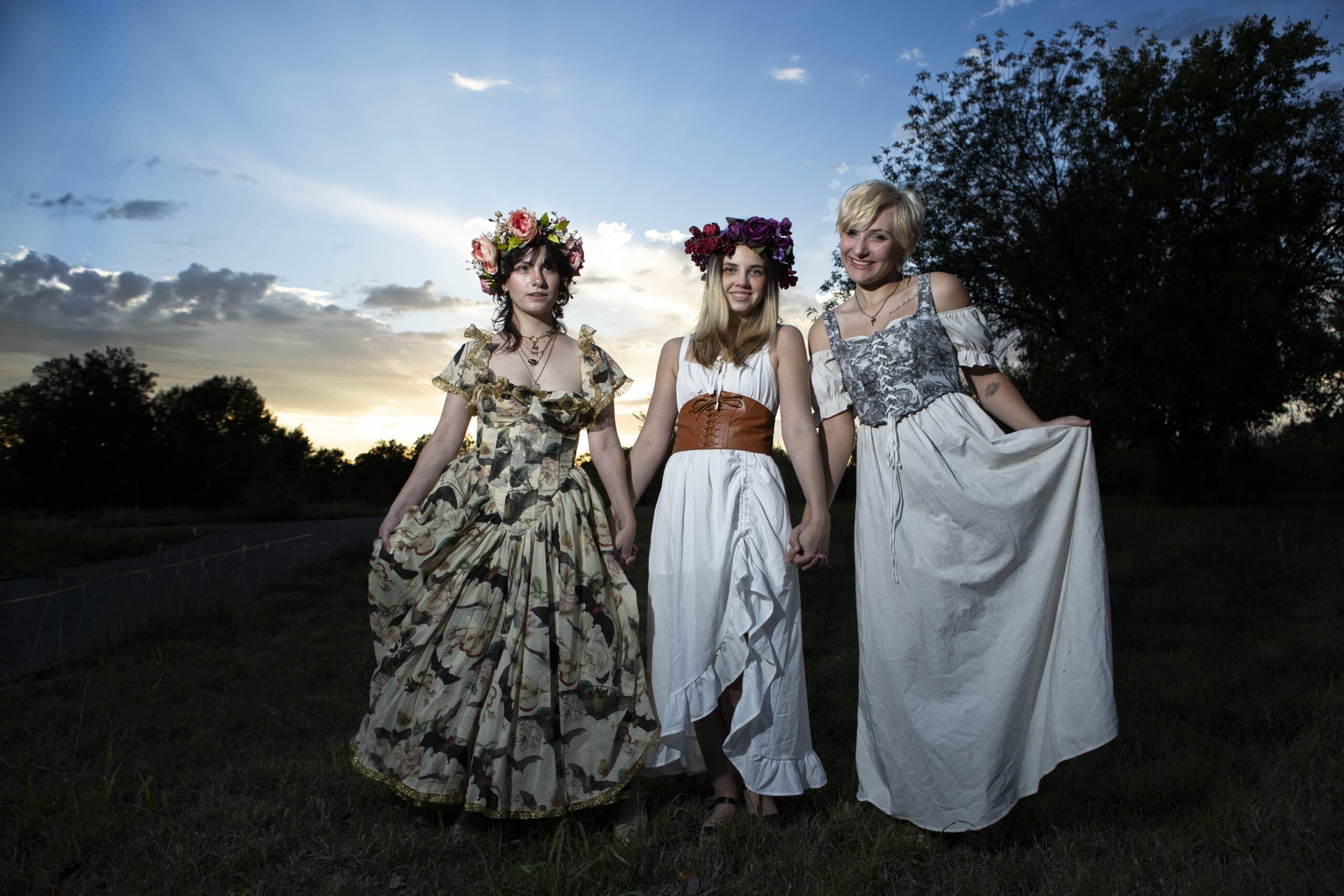 Three models pose together at sunset in Ray Trent Park. @maddenchaotic_cos wears a floral gown and pink flower crown, an unidentified model wears a white dress with a brown corset and purple flower crown, and @eternalhallowcos wears a flowing white and gray dress.
