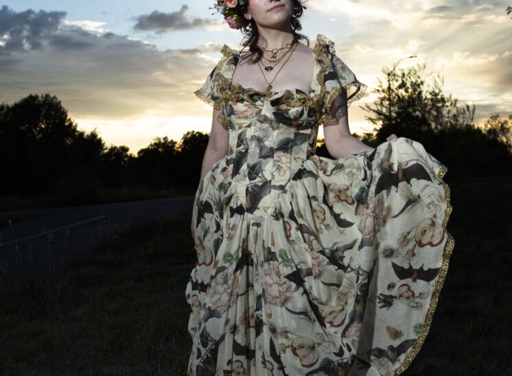 Model @maddenchaotic_cos wearing an ornate botanical-print gown with gold trim and a pink floral crown, gracefully holding her skirt against a golden sunset sky at Ray Trent Park in Midwest City, Oklahoma.