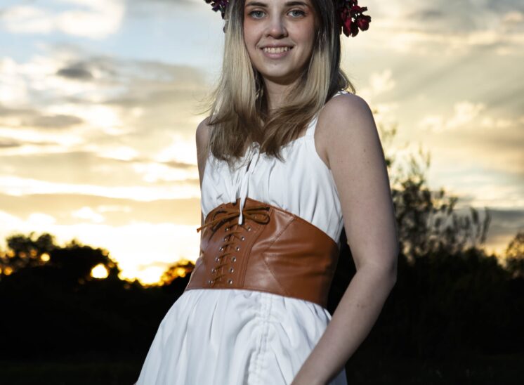 Model wearing a white dress, brown lace-up corset, and purple floral crown against a golden sunset at Ray Trent Park.