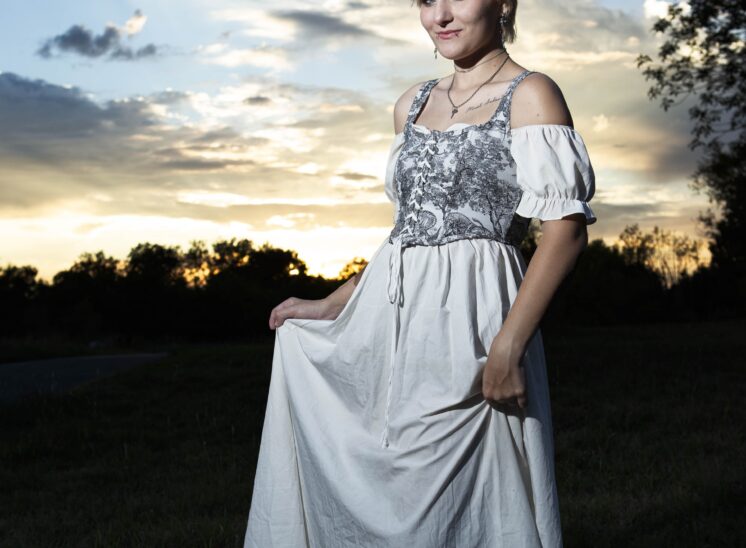 Model @eternalhallowcos wearing a flowing white dress with a gray patterned bodice, posing beneath a dramatic sunset sky at Ray Trent Park in Midwest City, Oklahoma.