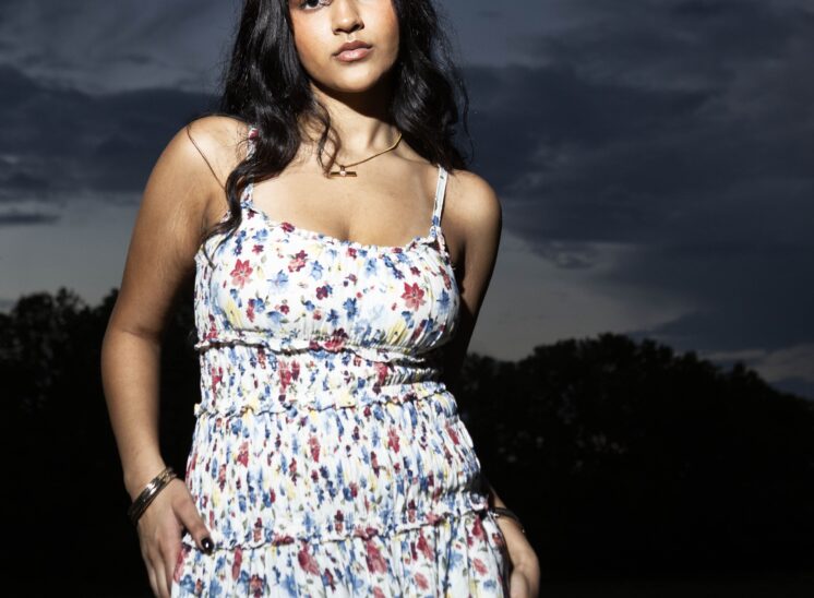 Model @alaya.jaffery wearing a flowing white floral sundress, posing beneath dramatic dark clouds at twilight in Ray Trent Park, Midwest City, Oklahoma.