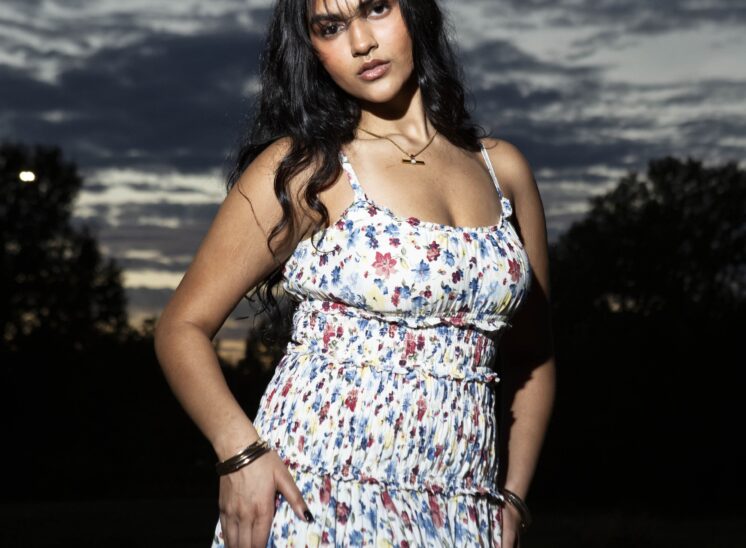 Model @alaya.jaffery wearing a white floral sundress, posing confidently with one hand on her hip beneath a dramatic, cloud-filled evening sky at Ray Trent Park.